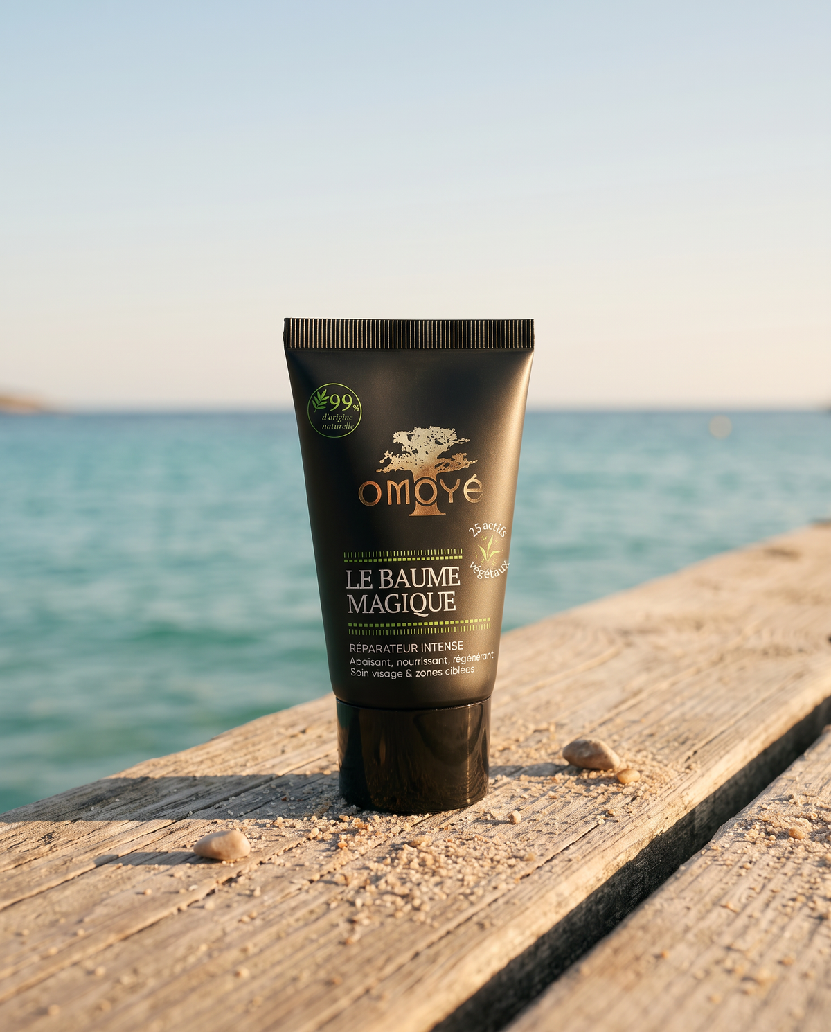 Tube du Baume Magique Omoyé posé sur un ponton en bois devant la mer turquoise, baume réparateur intense bio