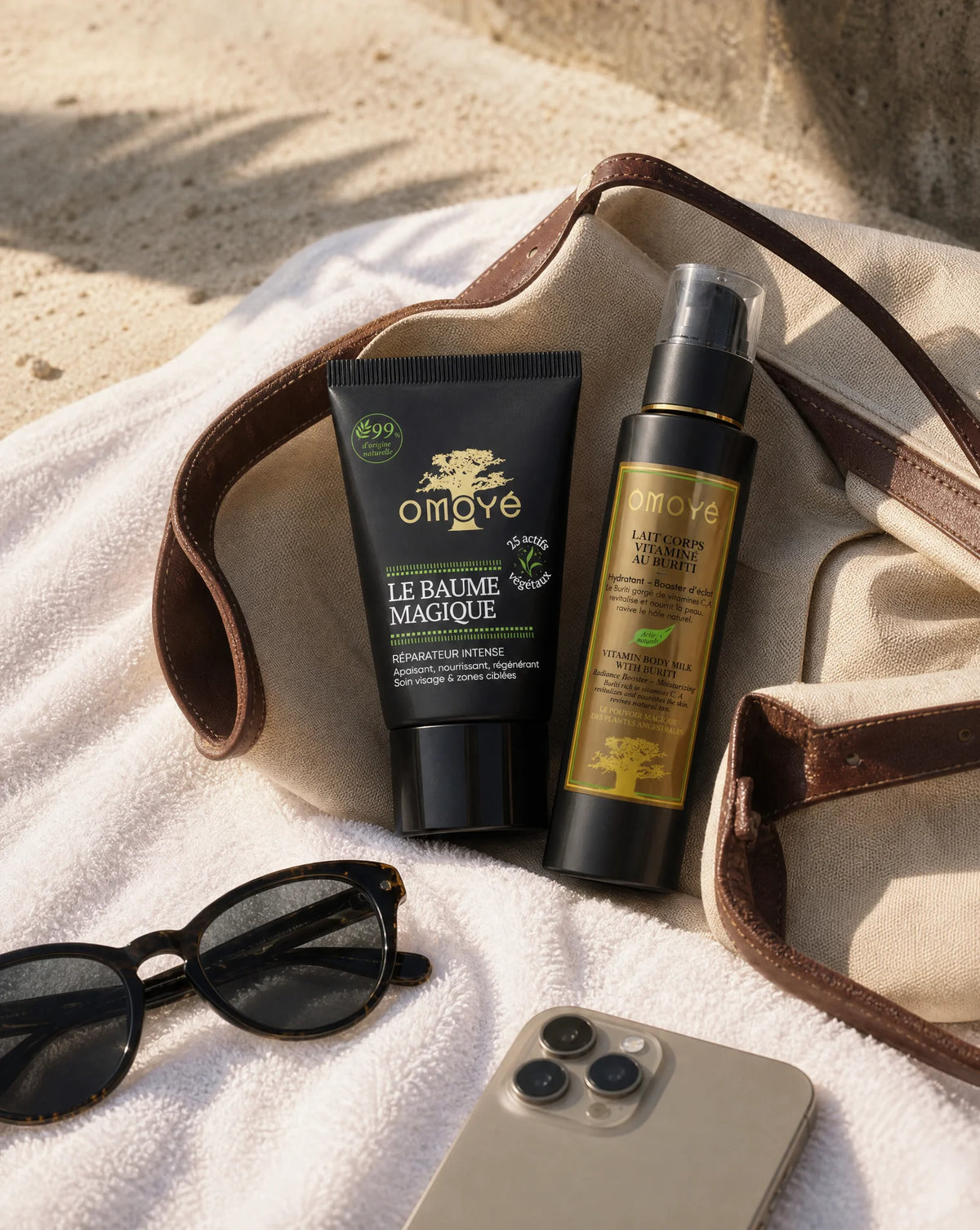 Le Baume Magique et le Lait Corps Vitaminé au Buriti posés sur une serviette de plage, à côté de lunettes de soleil, d'un téléphone et d'un sac en toile