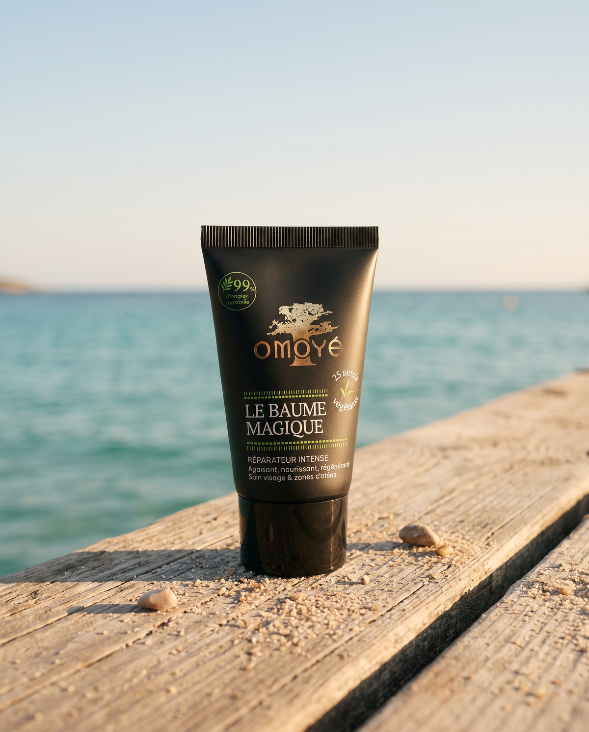 Tube du Baume Magique Omoyé posé sur un ponton en bois devant la mer turquoise, baume réparateur intense bio