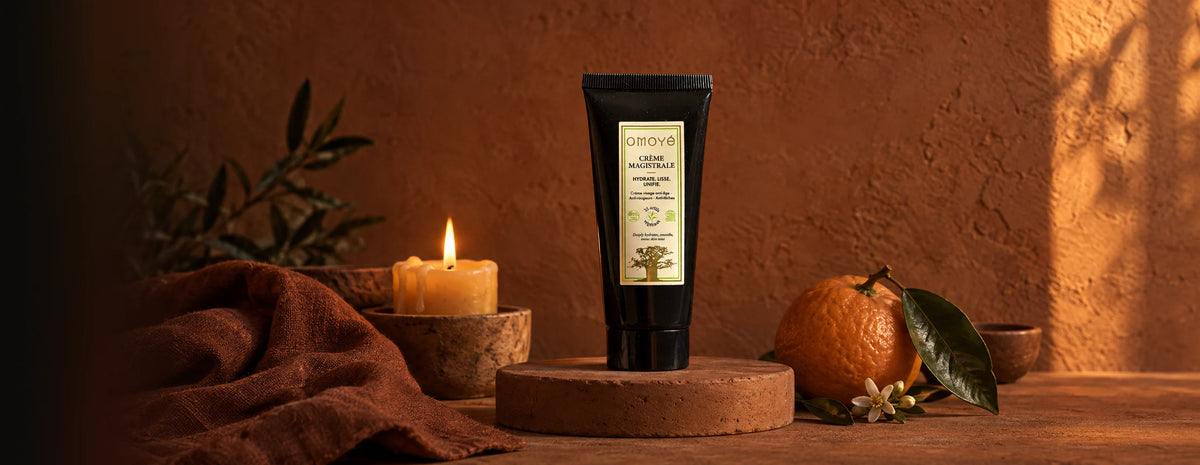 La Crème Magistrale OMOYÉ sur un socle en terre cuite, entre une bougie allumée et une orange amère en fleur, dans la lumière du soir