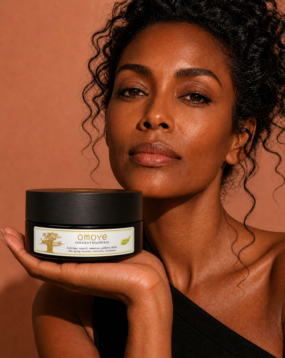 Femme tenant le pot de l'Onguent Magistral Omoyé sur fond terracotta, soin corps au jojoba